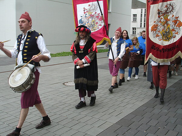 Gautschmeister 2015 (Foto: HTWK Leipzig) In mittelalterlicher Zunft läuft eine Schlange von Menschen über einen gepflasterten Weg. Viele von ihnen halten große Stoffbanner, auf denen Wappen aufgedruckt sind.