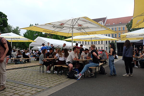 Eine Gruppe von Studierenden sitzt auf Bierbänken unter einem Sonnenschirm und unterhält sich. Im Hintergrund sind weitere Besucher des Hochschulsommerfestes zu sehen.