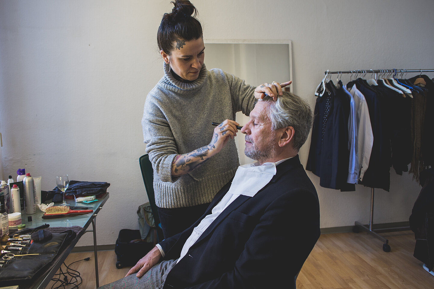 Ab in die Maske: Norbert Hippler (Foto: familie redlich AG/Niklas Schader) Norbert Hippler sitzt mit geschlossenen Augen auf einem Stuhl, während sein Gesicht von einer Visagistin bearbeitet wird.