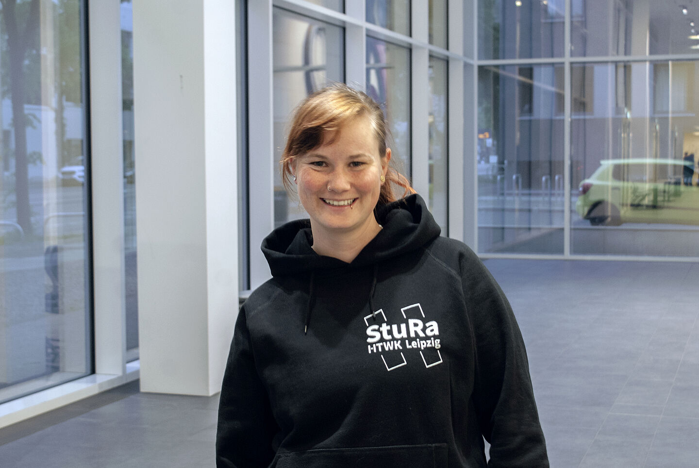 Maria trägt einen schwarzen Pullover mit weißem Aufdruck des StuRa-Logos, während sie im Foyer des Nieper-Baus in die Kamera lächelt