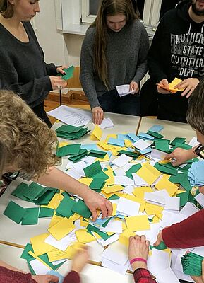 Eine Hand voll junger Menschen steht um einen Tisch herum und sortieren konzentriert Stimmzettel in vier verschiedenen Farben, die durchmischt auf dem Tisch liegen.