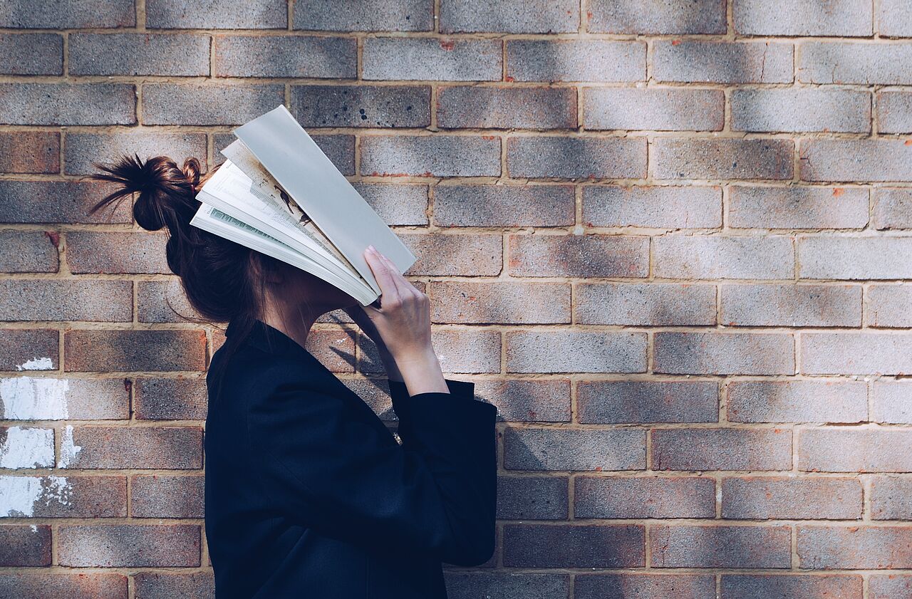 Eine Frau schlägt ein Buch vor ihr Gesicht, während sie vor einer Hauswand steht,